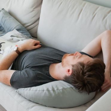 mężczyna leżący na kanapie, głowę wspartą ma na poduszce do medytacji i relaksacji w kształcie księżyca, z jasno szarego lnu.